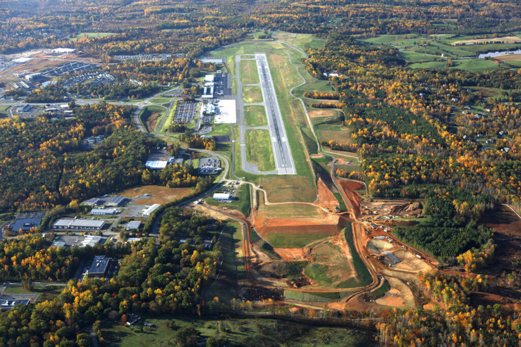 Vista aérea de un aeropuerto de una sola pista, rodeado de árboles y edificios dispersos.